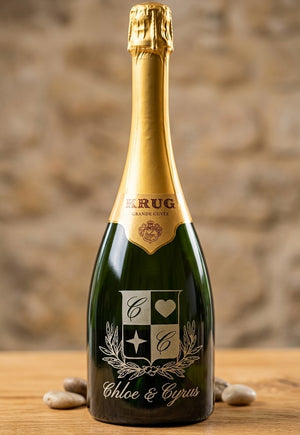 A bottle of Krug Grand Cru Champagne Cuvée Signature, personalized with a monogram and the names 'Chloe & Cyrus' on the label, set on a wooden surface with decorative stones against a rustic stone wall background.